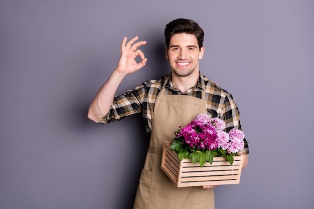 Photo photo of cheerful positive toothy beaming man smiling toothily showing ok sign holding flowers box with hand isolated grey color wall