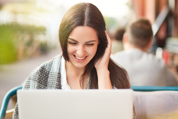 Photo of cheerful satisfied Caucasian woman looks positively at screen of laptop computer