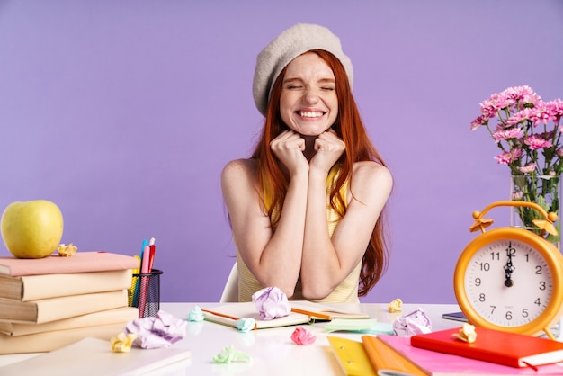 Photo of cheerful student girl sitting at desk with exercise books while doing homework isolated over purple wall