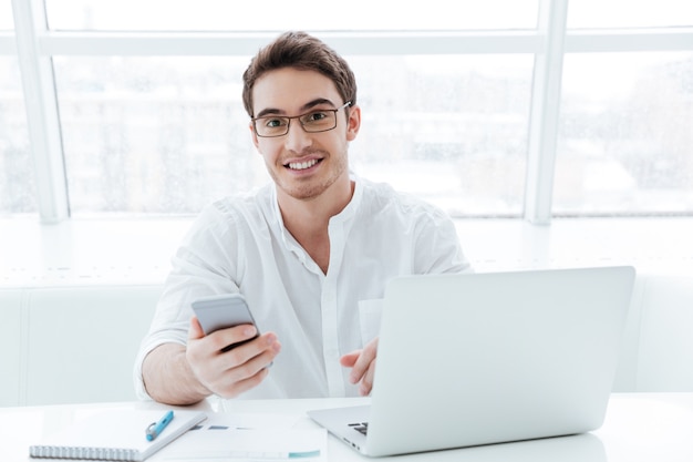 Photo of cheerful young man dressed in white shirt using laptop computer while chatting. Look at camera.