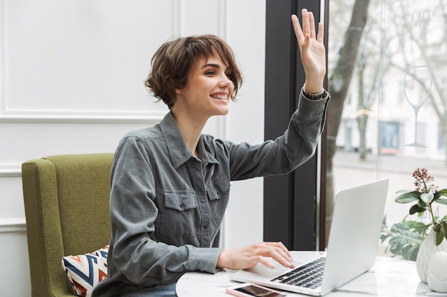 Photo of a cheerful young pretty business student woman sitting in cafe indoors using laptop computer coworking waving to friends.