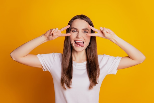 Photo of childish lady two v-signs cover eyes show tongue wear white t-shirt posing on yellow background