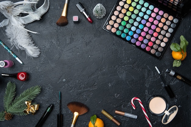 Photo of Christmas tree, mandarin, brushes, lipstick, palette with shadows on black table with space for text in center