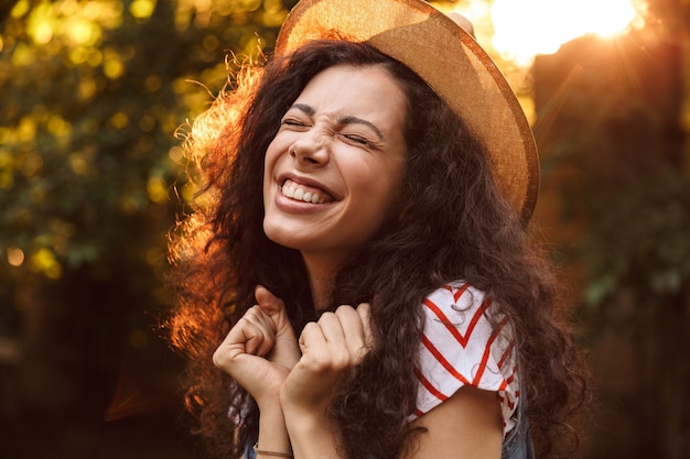 Photo closeup of brunette delighted woman wearing straw hat looking upward, while walking through park in sunny day