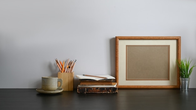 Photo of coffee cup, wooden pencil holder, olden book, pencil, picture frame and potted plant all of these are putting together on wooden black table with grey wall 