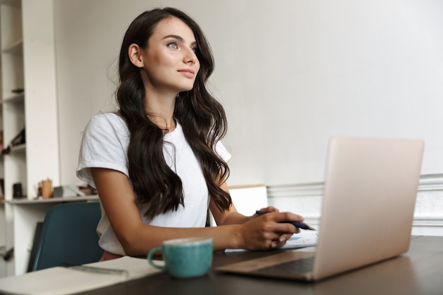 Photo of a concentrated young business woman indoors at home using laptop computer work with documents.