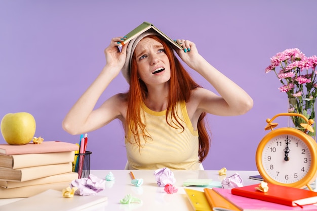 Photo of confused student girl holding exercise book on her head while doing homework isolated over purple wall