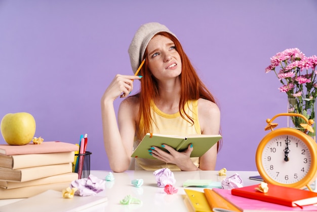 Photo of confused student girl writing in exercise book while doing homework isolated over purple wall