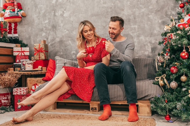 Photo of Cozy Happy Evening Young Couple at Home, Christmas Decor, Red Green Shades, Woman and man with gift box. New Year 2022 eve. Cup of tea or coffee