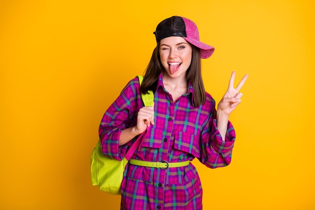 Photo of crazy optimistic teen girl show v-sign tongue out wear pink headwear dress green bag belt isolated on yellow color background