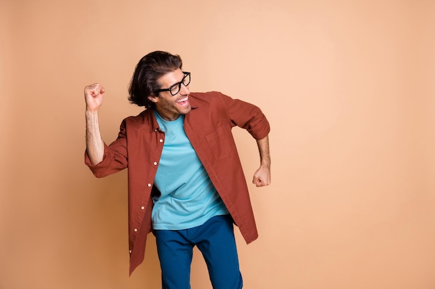 Photo of crazy young guy dance hold fists  look empty space wear specs brown t-shirt isolated beige color background