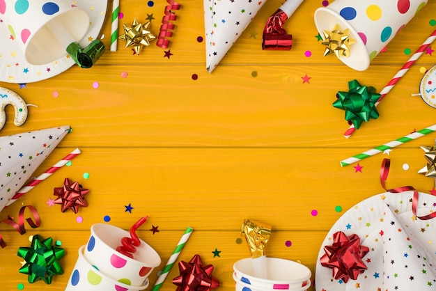 Above photo of cups drink plates confetti candle birthday hat bows party tubules plates and whistle isolated on the yellow wooden background with empty space