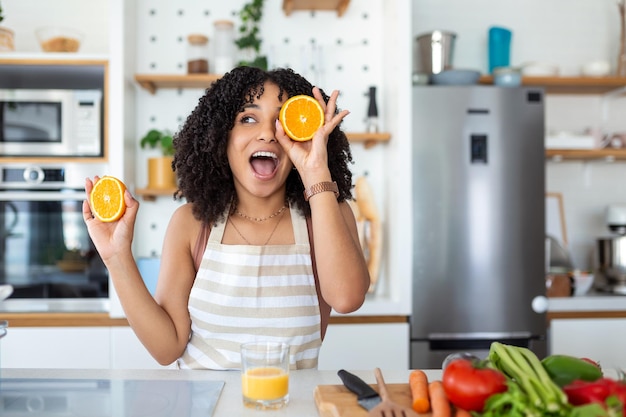 Photo of cute African American woman smiling and holding two orange parts while cooking vegetable salad in kitchen interior at home