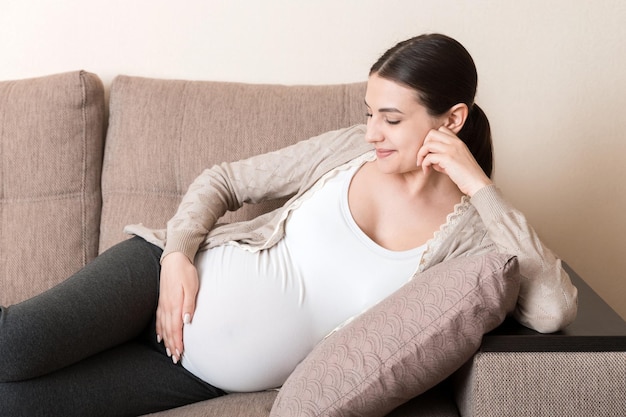 Photo of cute caucasian future mom lying in light living room on sofa and holding hands on tummy