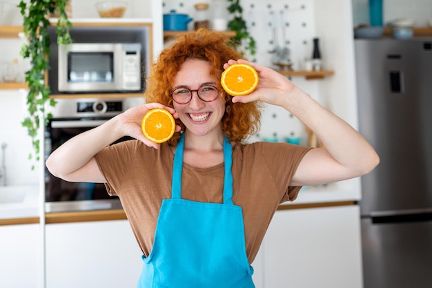 Photo of cute caucasian woman smiling and holding two orange parts while cooking vegetable salad in kitchen interior at home