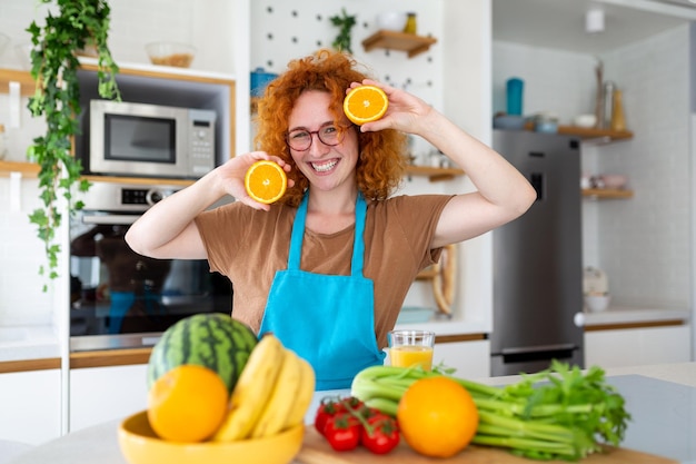 Photo of cute caucasian woman smiling and holding two orange parts while cooking vegetable salad in kitchen interior at home