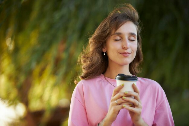 Photo of cute charming young lady in pink t-shirt,in park, with smile holding paper coffee cup.