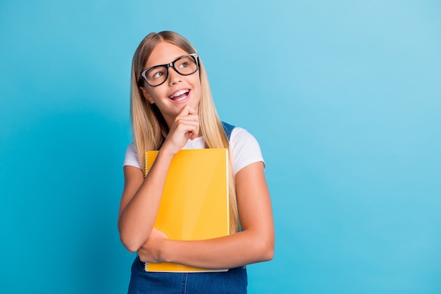 Photo of cute cheerful pupil girl thinking looking empty space wear spectacles hold book isolated on pastel blue color background