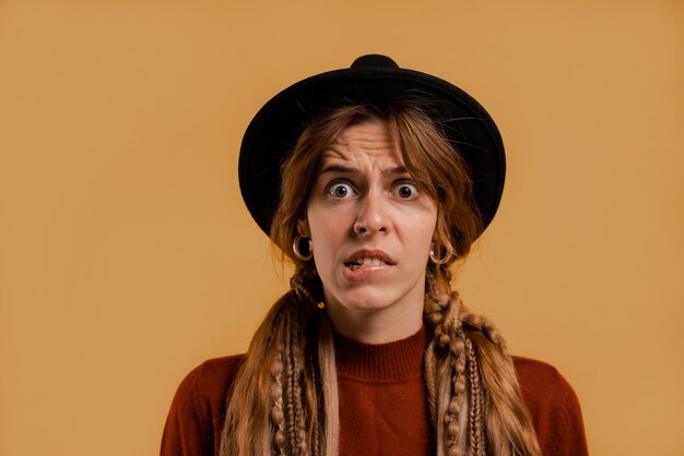 Photo of cute female farmer looks nervous about something. White woman wears denim overall and hat isolated brown color background.