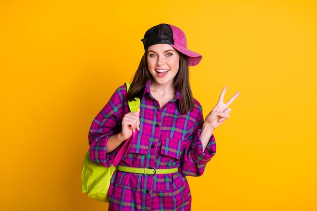 Photo of cute optimistic teen girl show v-sign wear pink headwear dress green bag belt isolated on yellow color background