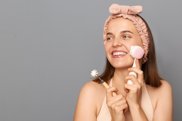 Photo of delighted cheerful woman holding items for makeup looking away with toothy smile copy space for advertisement empty area for promotional text