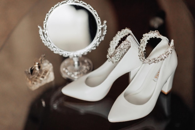 Photo of details at the wedding White chic shoes with decoration on the foot a beautiful mirror with a silver frame and perfume stand on a glass stand Shadows and light