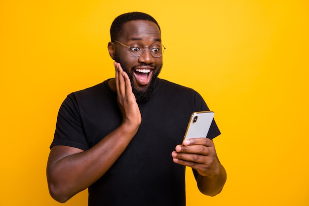 Photo of ecstatic overjoyed man browsing through his telephone rejoicing in receiving positive notifications in t-shirt isolated vivid color yellow wall 