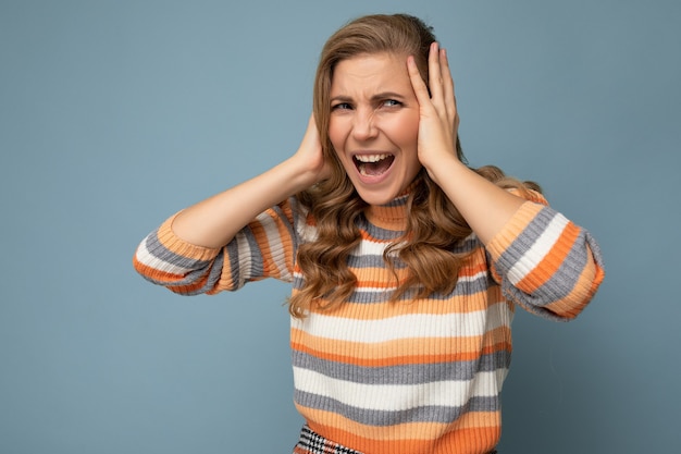 Photo of emotional angry young attractive blonde curly woman with sincere emotions wearing casual striped pullover isolated on blue background with copy space and covering ears trying not to hear and