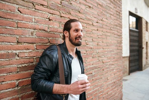 Photo of european unshaved man with tied hair standing over brick wall on city street and drinking takeaway coffee