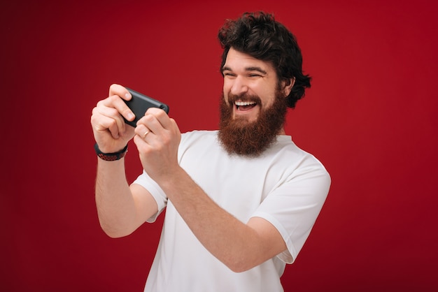 Photo of excited bearded man dressed in tshirt playing at phone over red wall