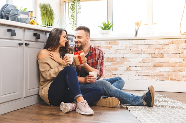 Photo of excited brunette couple man and woman 20s having breakfast in apartment while sitting on the floor with present box. Celebrating holiday.