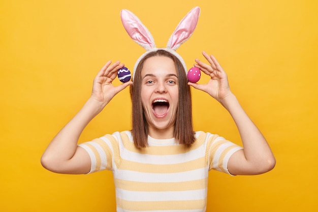 Photo of excited joyful cheerful woman wearing rabbit ears holding colorful Easter eggs having fun while celebrating yelling with happy face isolated on yellow background