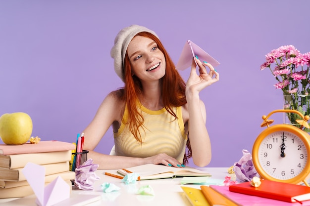 Photo of excited student girl holding paper plane while doing homework at desk isolated over purple wall