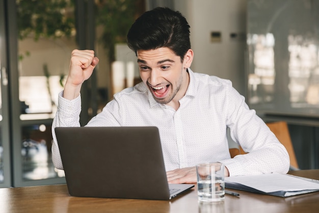 Photo of excited young man 30s wearing white shirt and bluetooth earbuds screaming and clenching fist like winner, while looking at laptop in office