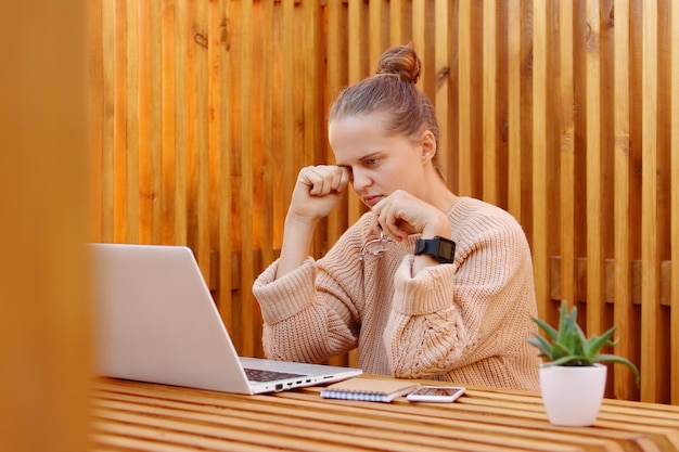 Photo of exhausted attractive young adult woman with bun hairstyle wearing beige sweater working on laptop against wooden wall being overworked rubbing eyes fells pain in eyes