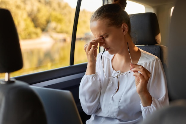 Photo of exhausted depressed woman wearing white shirt sitting in car with notebook being exhausted after hard working day suffering headache and eyes hurt keeps eyes closed