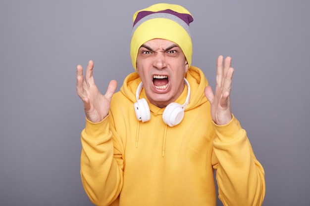 Photo of extremely angry crazy young adult man wearing beanie hat and yellow casual hoodie standing isolated over gray background screaming with aggression raised his arms has quarrel