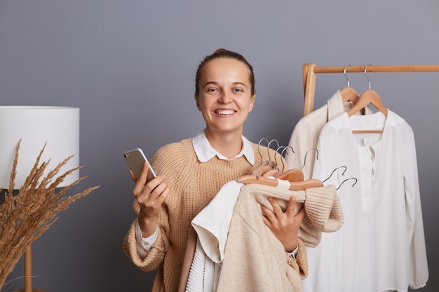 Photo of extremely happy woman shopper holding clothes on racks in modern boutique choosing new look or wants to change image holding smart phone checking fashion blog while doing shopping