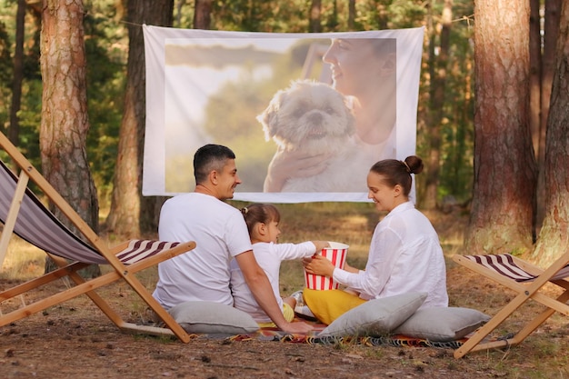 Photo of family sitting in the forest on deck chairs with their little daughter and watching movie on projector parents looking at each other with smile family enjoying time spending together