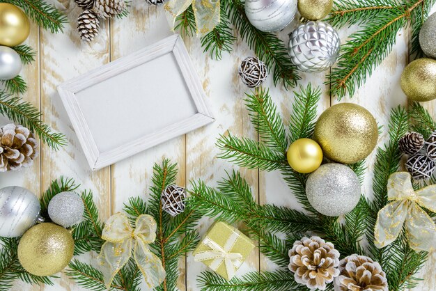 Photo frame between Christmas decoration, with golden color balls and pine cones on a white wooden table. Top view, frame to copy space.