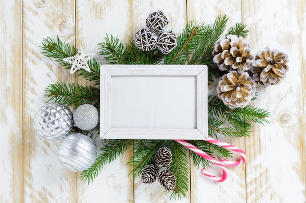 Photo frame between Christmas decoration, with white balls and pine cones on a white wooden table. Top view, frame to copy space