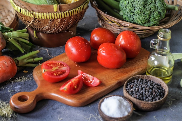 Photo of fresh tomatoes in cutting board, around vegetables, carrot, salt, black pepper, corn, broccoli. Slice tomatoes. Harvesting tomatoes. Wooden table.