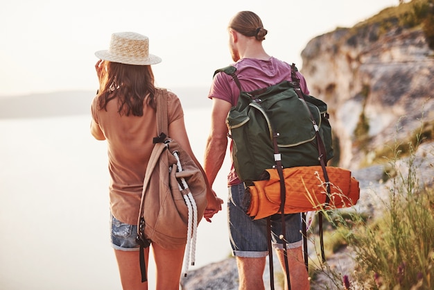 Photo from the back. Young couple have decided to spend their holiday in active way on the edge of the gorgeous rock with lake at background.