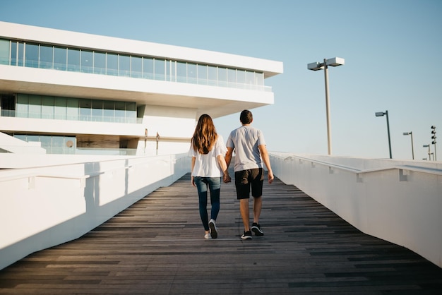 A photo from behind of a Hispanic man and brunette woman who are strolling in the harbor in Spain