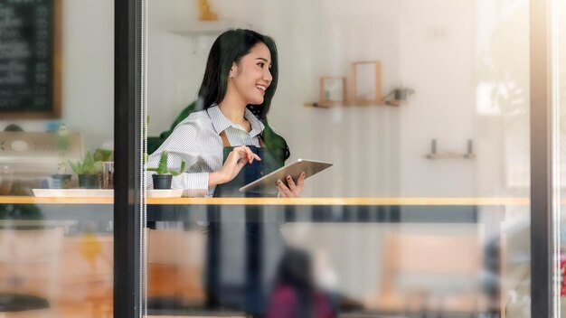 Photo from outside the glass side view of a small business owner a happy Asian woman sitting at the counter holding a tablet at a cafÃ©.