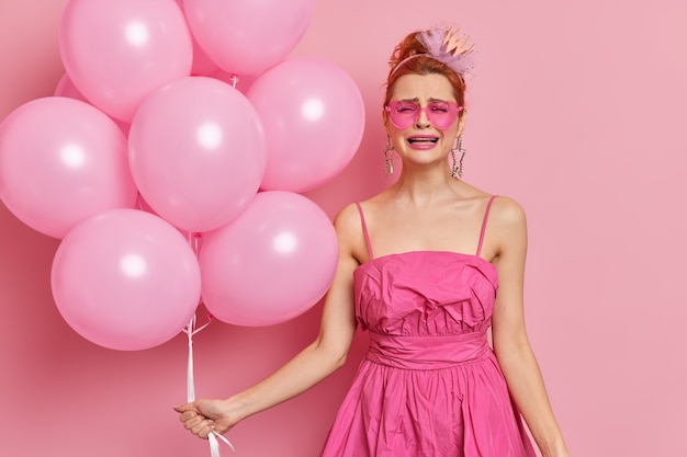 Photo of frustrated redhead woman cries from despair poses in festive clothing with bunch of inflated balloons isolated over pink wall. Bad holiday concept. This party sucks. Human emotions