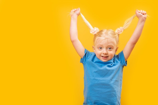 Photo of funny little girl in blue tshirt having fun and holding two ponytails with her blonde hair Yellow background Copy space