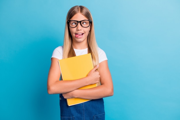 Photo of funny pupil girl do not want do homework show tongue wear spectacles hold book isolated on pastel blue color background