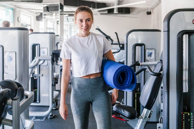 Photo of A girl with a gymnastic rug in her hands poses on the background of the gym. Smiling and looking at the camera