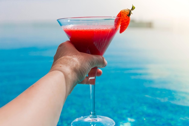 Photo of a glass of strawberry cocktail in female hands swimming pool